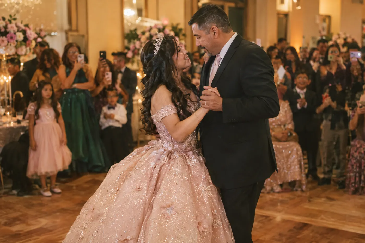 A quinceañera in a sparkling ball gown dancing with her father as family members film and take photos