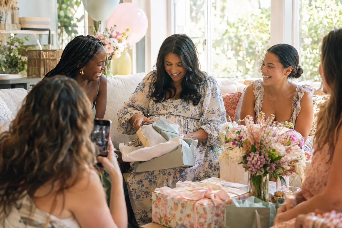 Friends laughing as a mother-to-be opens a gift at a bright, flower-filled baby shower