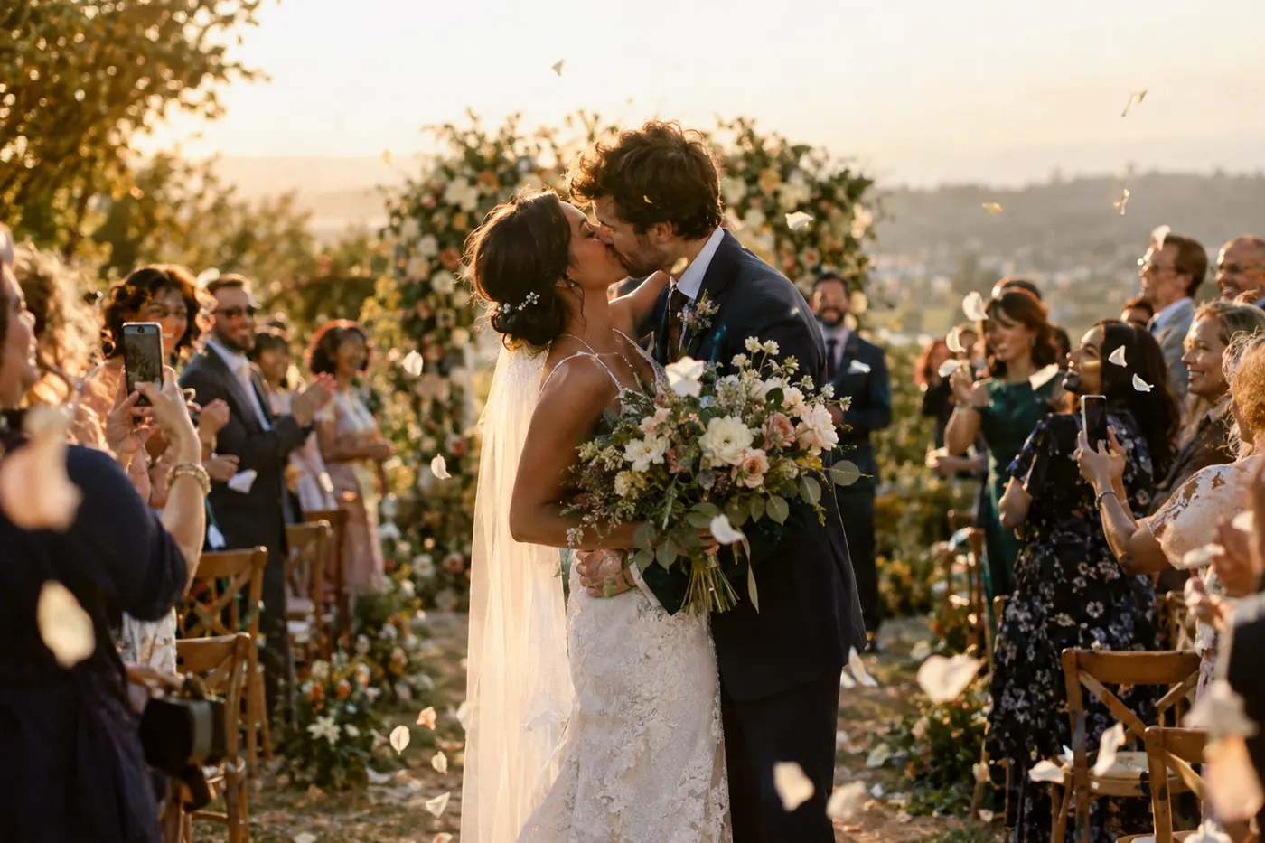 Wedding guests cheering and taking photos with their phones as the couple kisses at an outdoor golden-hour ceremony